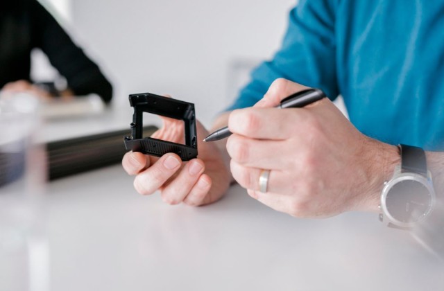 Ein Mann hält ein gefrästest schwarzbeschichtetes CNC Fertigungsbauteil inder Hand und zeigt mit einem Kugelschreiber auf eine Kante des Bauteils im Hntergrund sitzt eine weitere Person und lässt sich von dem Mann beraten