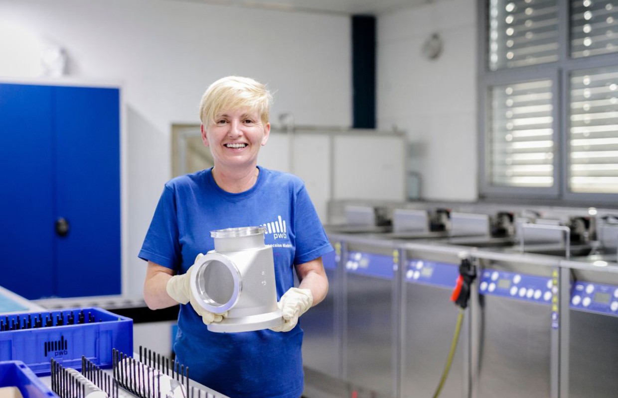 Blonde Frau mit blauem Shirt hält ein CNC gefertigtes Bauteil in den Händen in einem Reinigungsraum