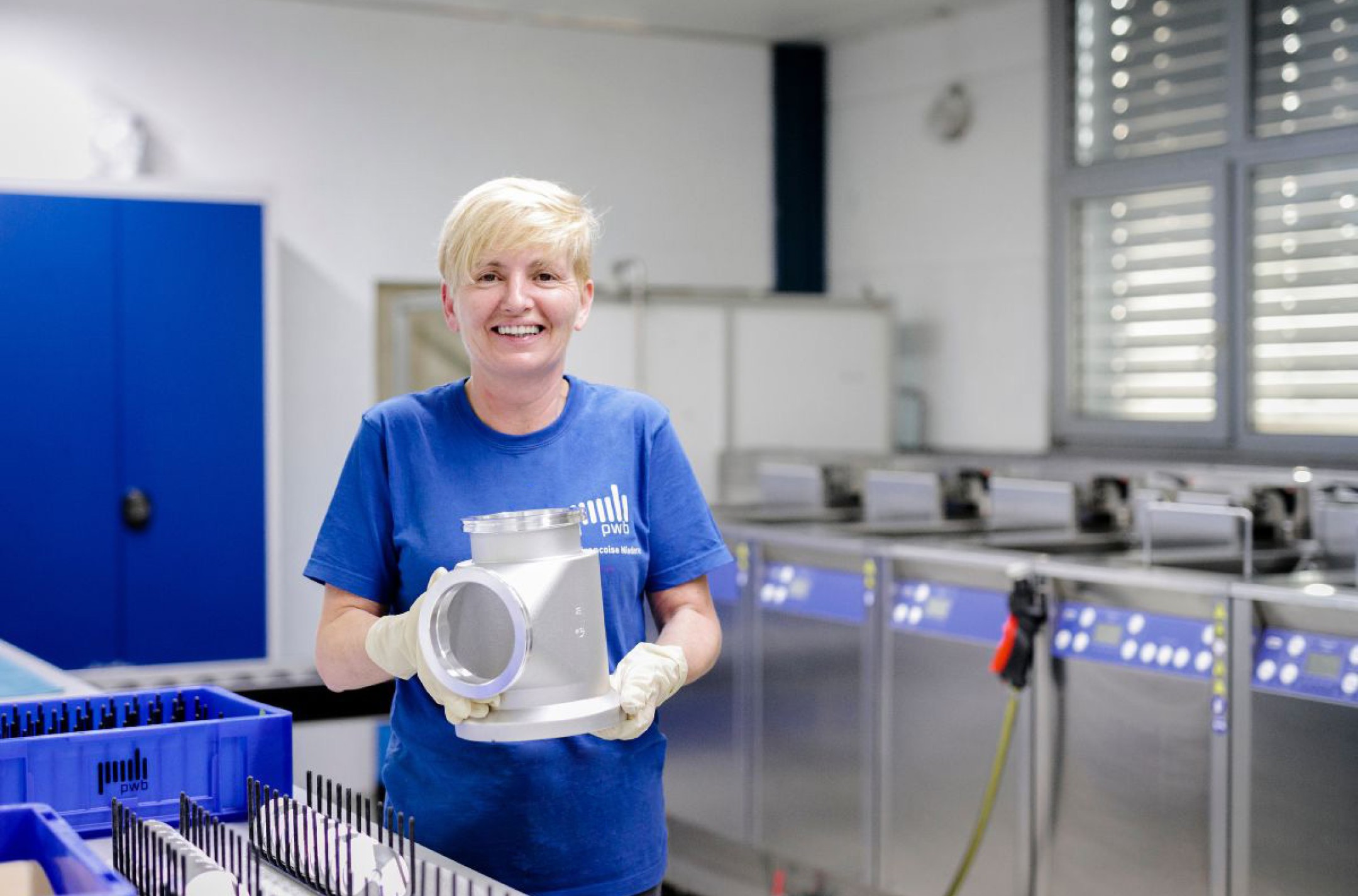 Blonde Frau mit blauem Shirt hält ein CNC gefertigtes Bauteil in den Händen in einem Reinigungsraum